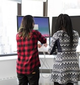 two women standing in front of two black monitors